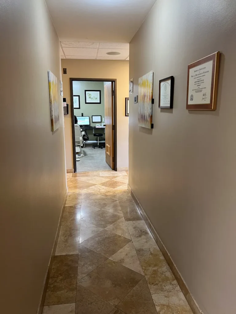 Hallway with beige walls, framed certificates, diamond-pattern tile floor, and open doorway to operatory
