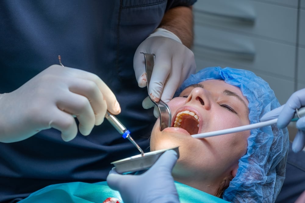 Oral Surgeon works on patient mouth during application of bone graft material. Assistant helps maintain access while woman lying on chair