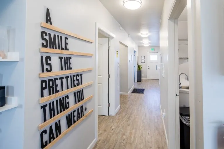 Hallway with wood-look flooring; wall sign reads “A SMILE IS THE PRETTIEST THING YOU CAN WEAR” in black block letters on wood slats