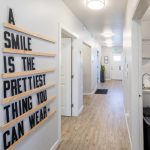 Hallway with wood-look flooring; wall sign reads “A SMILE IS THE PRETTIEST THING YOU CAN WEAR” in black block letters on wood slats