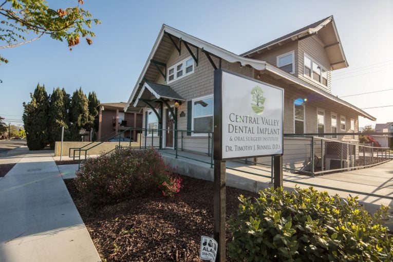 Exterior: “Central Valley Dental Group” sign on post, beige building with gabled roof, shrubs, and clear sky