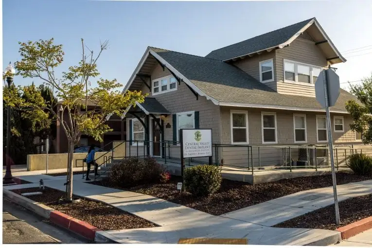 Single-story dental office with gray siding, gabled roof, front porch, and landscaped walkway under sunny sky