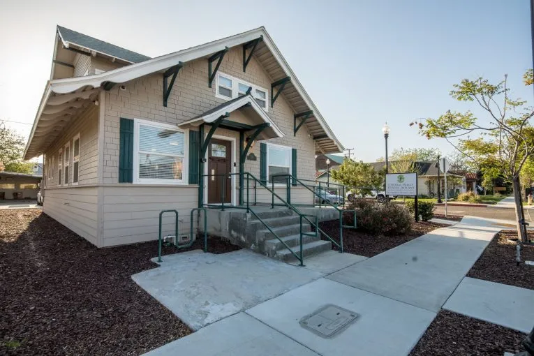 Front entrance: single-story building with covered porch, glass door, green ramp, and landscaped walkway