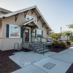 Front entrance: single-story building with covered porch, glass door, green ramp, and landscaped walkway
