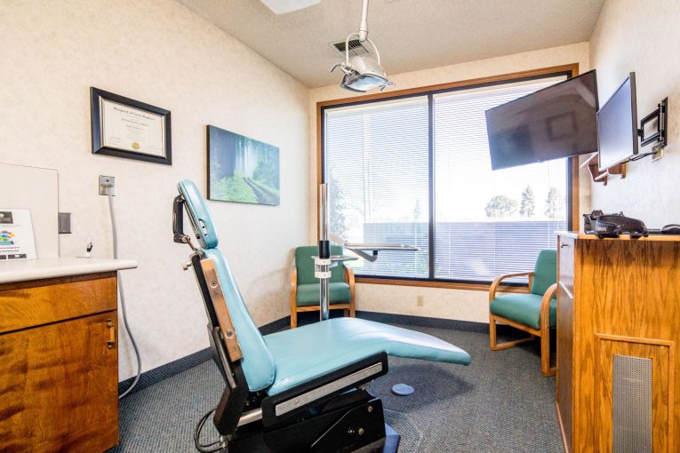 Operatory with teal dental chair, large window, wood cabinets, and wall art
