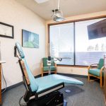 Operatory with teal dental chair, large window, wood cabinets, and wall art