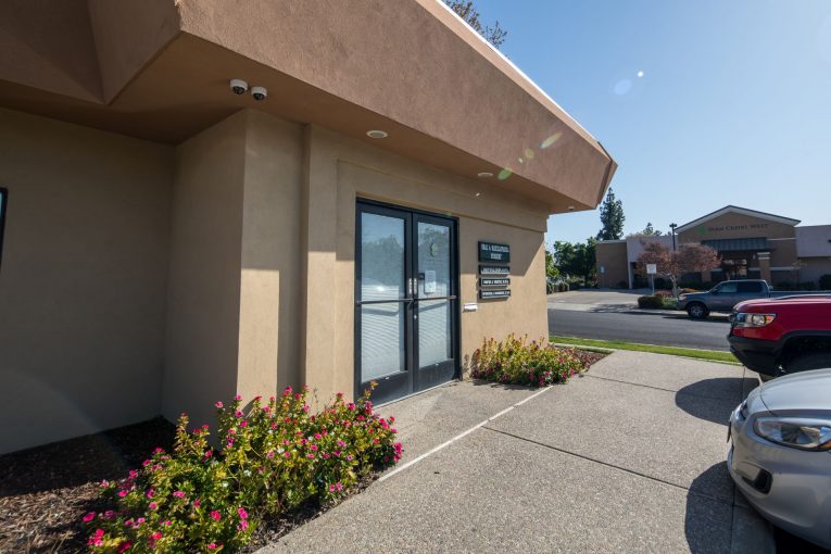 Office entrance: beige building, glass doors, flower bed, parked silver car