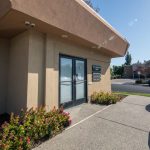 Office entrance: beige building, glass doors, flower bed, parked silver car