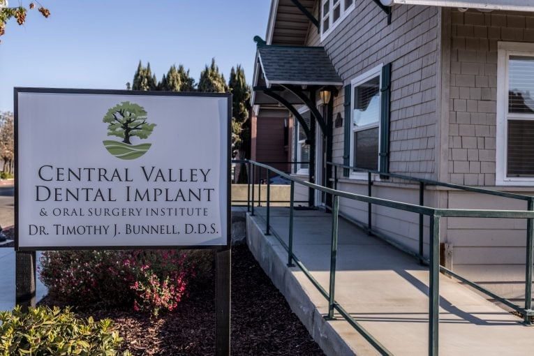 Close-up of practice sign on post: “Central Valley Dental Implant & Oral Surgery Institute, Dr. Timothy J. Rennell, D.D.S.” with tree logo; ADA-accessible ramp nearby