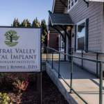 Close-up of practice sign on post: “Central Valley Dental Implant & Oral Surgery Institute, Dr. Timothy J. Rennell, D.D.S.” with tree logo; ADA-accessible ramp nearby