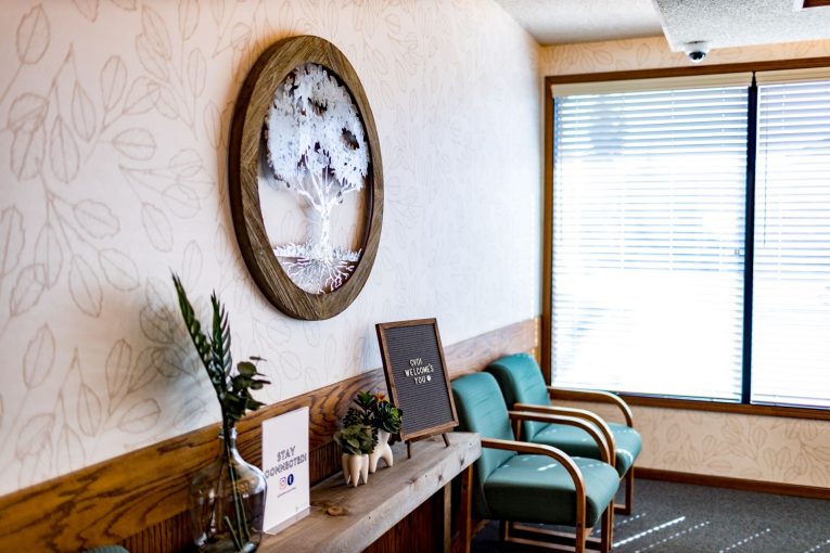 Seating nook: two teal chairs, wooden console, oval wall art, window with blinds.