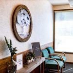 Seating nook: two teal chairs, wooden console, oval wall art, window with blinds.