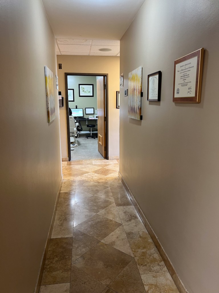 Hallway with beige walls, framed certificates, diamond-pattern tile floor, and open doorway to operatory