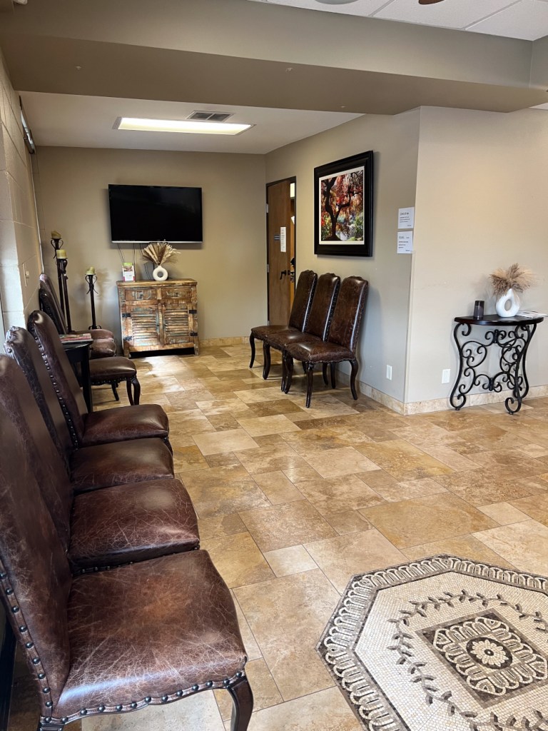 Waiting room with row of brown leather chairs, tiled floor, patterned rug, and wall-mounted TV at far end