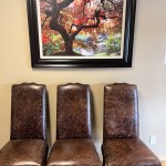 Three brown leather chairs aligned beneath framed landscape painting (autumn trees, river scene)