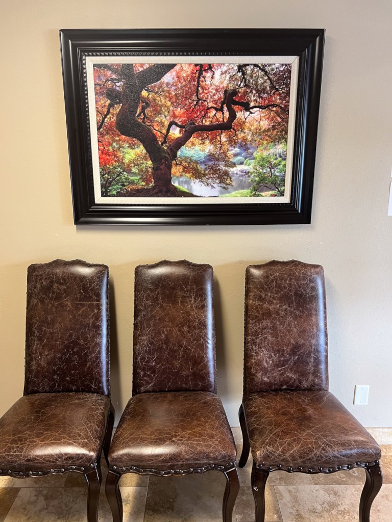 Three brown leather chairs aligned beneath framed landscape painting (autumn trees, river scene)